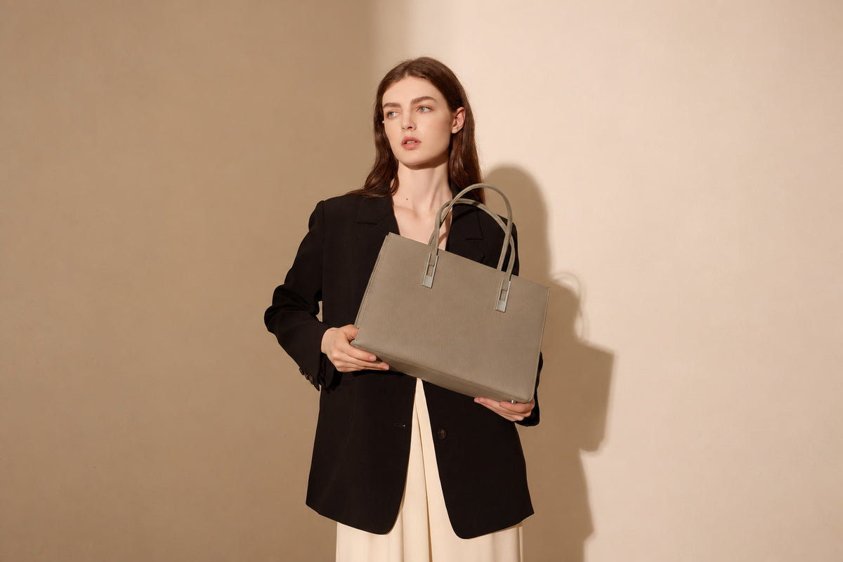 Sac Melinda taupe tenu devant soi, photographié dans un décor beige élargi