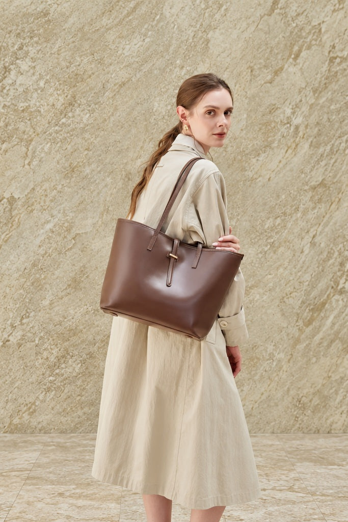 Cabas Marina chocolat porté à l’épaule avec un manteau beige, devant un mur en pierre